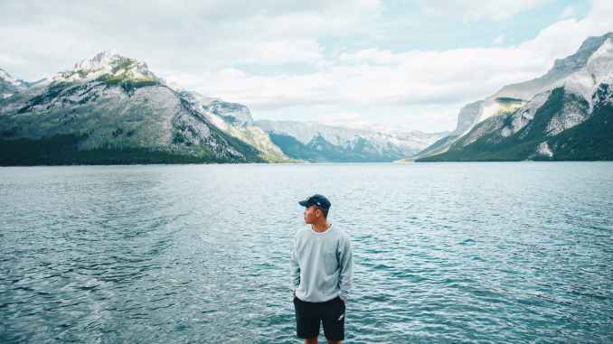 a man in gray sweater standing near the body of water while looking at the mountain